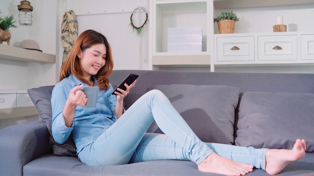 Mujer asiática sonriente atractiva hermosa que usa el smartphone que sostiene una taza de café caliente