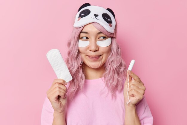 Una mujer asiática sonriente de cabello rosado complacida tiene una expresión de ensueño aplica parches de belleza debajo de los ojos para refrescar la piel elige el mejor producto de higiene sostiene una toalla sanitaria y un tampón tiene menstruación