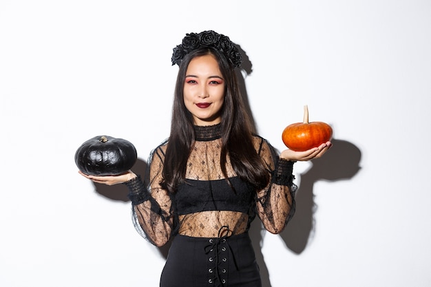 Mujer asiática sonriente complacida celebrando halloween, vistiendo traje de bruja, sosteniendo calabazas