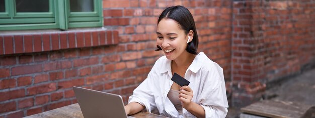 Mujer asiática sonriente con computadora portátil y auriculares inalámbricos pagando con tarjeta de crédito comprando en línea sentado