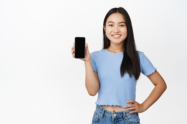 Mujer asiática sonriente confiada que muestra la pantalla del teléfono móvil cogidos de la mano en la cintura de pie en camiseta sobre fondo blanco