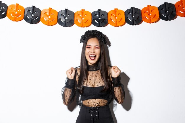 Mujer asiática sonriente despreocupada en traje de bruja disfrutando de la fiesta de halloween, bailando y regocijándose, de pie sobre fondo blanco con decoración de serpentinas de calabaza.