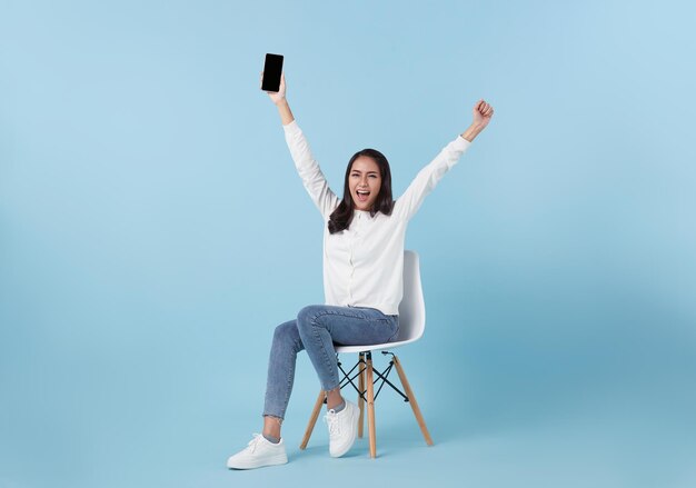 Mujer asiática sonriente feliz sosteniendo un teléfono inteligente sentado en una silla y ganando el premio