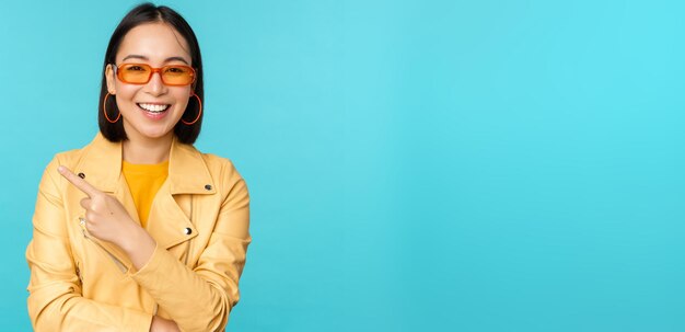 Mujer asiática sonriente con gafas de sol apuntando con el dedo a la izquierda mostrando el logotipo de la pancarta o el anuncio de pie sobre fondo azul Copiar espacio