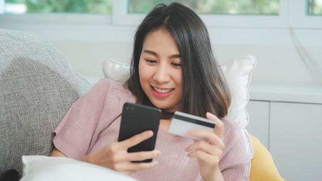 Mujer asiática sonriente joven que usa el smartphone que compra compras en línea con tarjeta de crédito mientras que miente en el sofá cuando se relaja en sala de estar en casa. Mujeres de origen étnico latino e hispano en el concepto de casa.