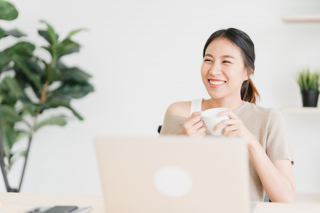Mujer asiática sonriente de los jóvenes hermosos que trabaja en el ordenador portátil y que bebe el café