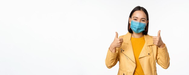 Mujer asiática sonriente con mascarilla médica que muestra el pulgar hacia arriba apoyando el uso de equipo de protección contra la pandemia de coronavirus covid19 de fondo blanco