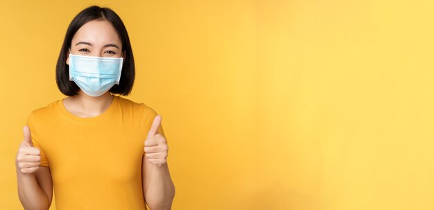 Mujer asiática sonriente con mascarilla médica que muestra el pulgar hacia arriba como aprobación y recomienda algo de pie