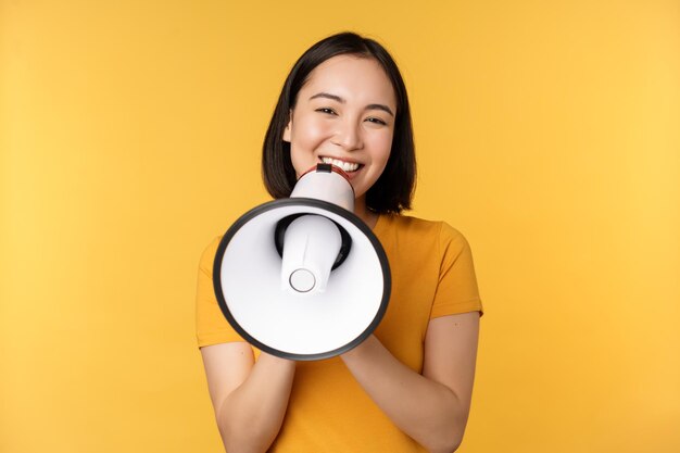Mujer asiática sonriente de pie con megáfono anunciando algo de producto publicitario de pie sobre fondo amarillo