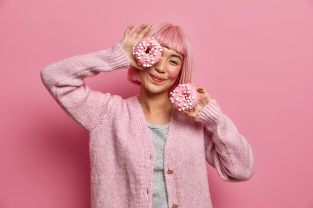 La mujer asiática sonriente positiva se divierte y sostiene dos deliciosas donas, juega con productos azucarados, disfruta de un apetitoso postre, viste un jersey informal,