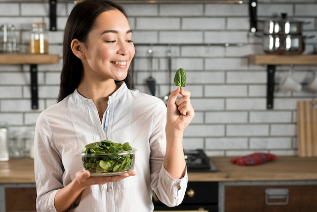 Mujer asiática sonriente que mira la hoja de la albahaca en cocina