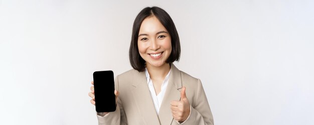 Mujer asiática sonriente que muestra la pantalla del teléfono inteligente y los pulgares hacia arriba La persona corporativa demuestra la interfaz de la aplicación del teléfono móvil de pie sobre fondo blanco