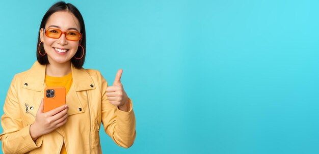 Mujer asiática sonriente que muestra los pulgares hacia arriba grabando en el teléfono móvil usando la aplicación de teléfono inteligente y recomendando
