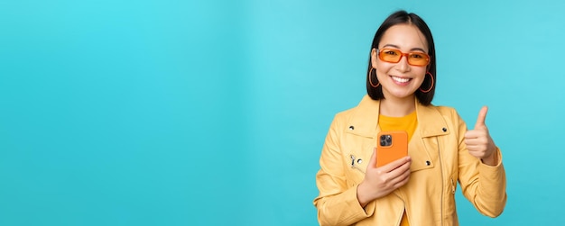 Mujer asiática sonriente que muestra los pulgares hacia arriba grabando en el teléfono móvil usando la aplicación de teléfono inteligente y recomendando