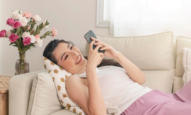 Una mujer asiática sonriente y relajada en el sofá de su casa está usando un teléfono inteligente y enviando mensajes de texto