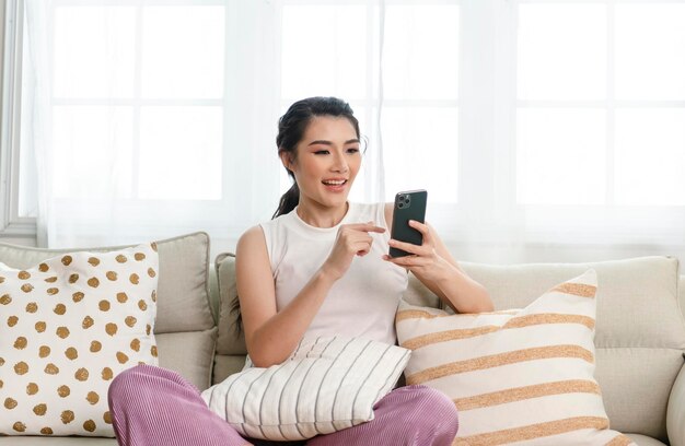 Una mujer asiática sonriente y relajada en el sofá de su casa está usando un teléfono inteligente y enviando mensajes de texto