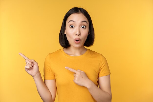 Mujer asiática sonriente señalando con el dedo a la izquierda mostrando un anuncio en un espacio de copia vacío de pie sobre un fondo amarillo