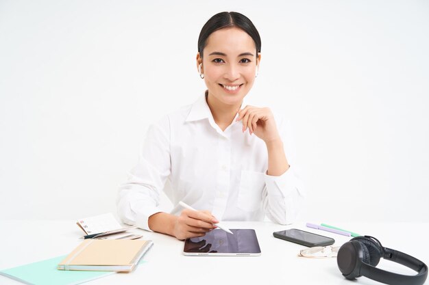 Foto gratuita una mujer asiática sonriente se sienta en su oficina, trabaja en una tableta digital, tiene auriculares, un teléfono inteligente y trabaja