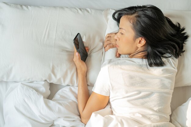 Mujer asiática con una sonrisa atractiva usa el teléfono inteligente tableta en la cama blanca. Vista superior