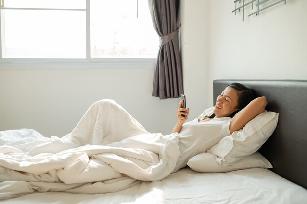 Mujer asiática con una sonrisa atractiva usa el teléfono inteligente tableta en la cama blanca.