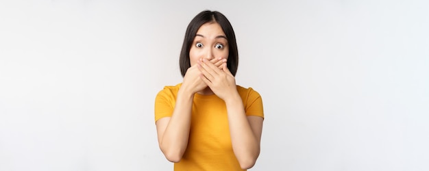 Una mujer asiática sorprendida cubre la boca con las manos mirando sorprendida con una expresión facial sin palabras de pie en una camiseta amarilla contra el fondo blanco