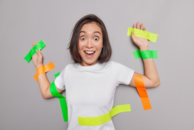 Mujer asiática sorprendida positiva pegada a la pared con cintas adhesivas de colores se siente feliz de no poder creer sus ojos vestidos con camiseta blanca aislada sobre pared gris