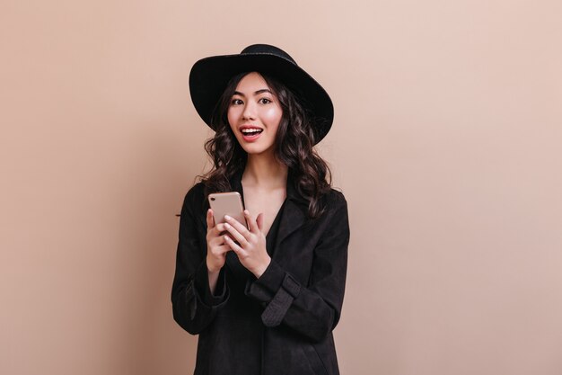 Mujer asiática sorprendida sosteniendo smartphone y mirando a cámara. Elegante mujer rizada en abrigo posando con gadget.