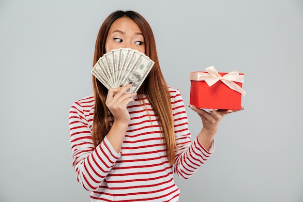 Mujer asiática sorprendida en suéter escondiéndose detrás de un dinero y mirando el regalo sobre fondo gris