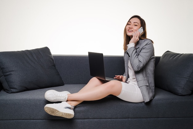 Mujer asiática en el teléfono usando la computadora portátil en casa en la sala de estar. Trabajando desde casa en cuarentena.