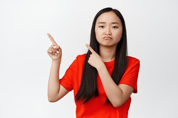 Mujer asiática triste y de mal humor señalando con el dedo en la esquina superior izquierda, haciendo pucheros y frunciendo el ceño molesto, de pie con una camiseta roja sobre blanco