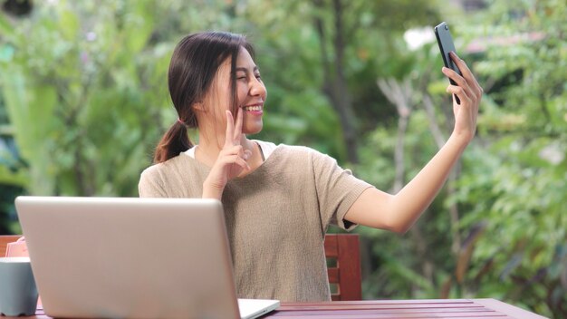 Mujer asiática utilizando teléfono móvil selfie post en las redes sociales, mujer relajarse sentirse feliz mostrando bolsas de compras sentado en la mesa en el jardín en la mañana.