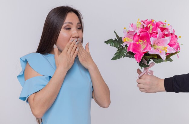 Mujer asiática en vestido azul mirando feliz y sorprendido cubriendo la boca con las manos mientras recibe ramo de flores