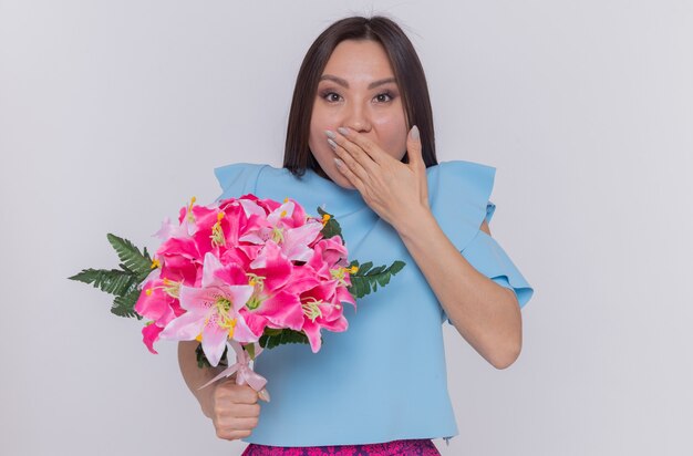 Mujer asiática en vestido azul con ramo de flores feliz y sorprendido cubriendo la boca