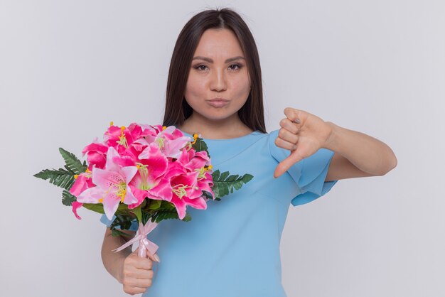Mujer asiática en vestido azul con ramo de flores mirando al frente disgustado mostrando el pulgar hacia abajo celebrando el día internacional de la mujer de pie sobre una pared blanca