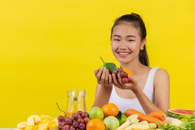 Una mujer asiática vistiendo una camiseta blanca. Ambas manos sostenían fruta y la mesa está llena de varias frutas.