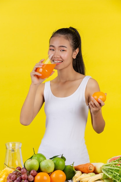 Una mujer asiática vistiendo una camiseta blanca. Beber zumo de naranja y sobre la mesa hay muchas frutas.