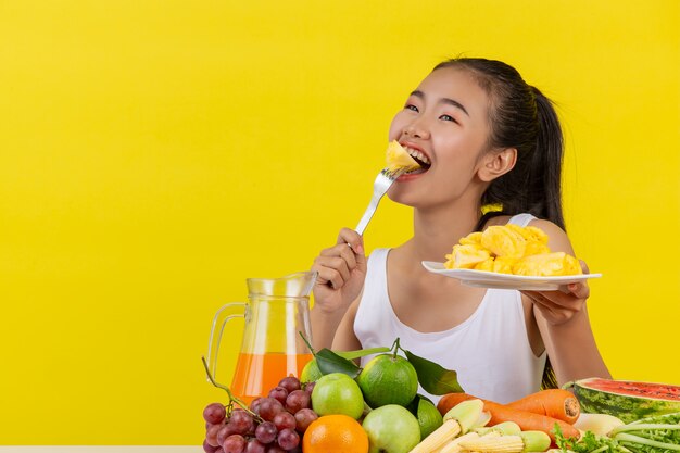 Una mujer asiática vistiendo una camiseta blanca. Comer piña Y la mesa está llena de varios tipos de frutas.