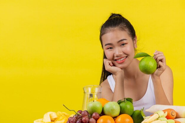 Una mujer asiática vistiendo una camiseta blanca. La mano izquierda sostiene unas naranjas verdes y la mesa está llena de muchos tipos de frutas.