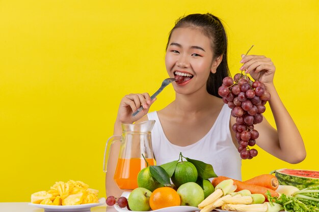 Una mujer asiática vistiendo una camiseta blanca. La mano izquierda sostiene un racimo de uvas. La mano derecha recoge las uvas para comer y la mesa está llena de varias frutas.