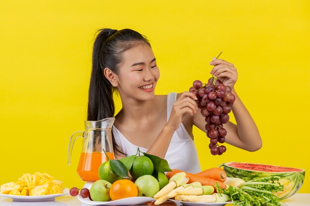 Una mujer asiática vistiendo una camiseta blanca. La mano izquierda sostiene un racimo de uvas. La mano derecha recoge las uvas para comer y la mesa está llena de varias frutas.