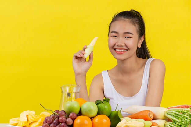 Una mujer asiática vistiendo una camiseta blanca. Sostén el elote con tu mano derecha. Y en la mesa hay muchas frutas diferentes.