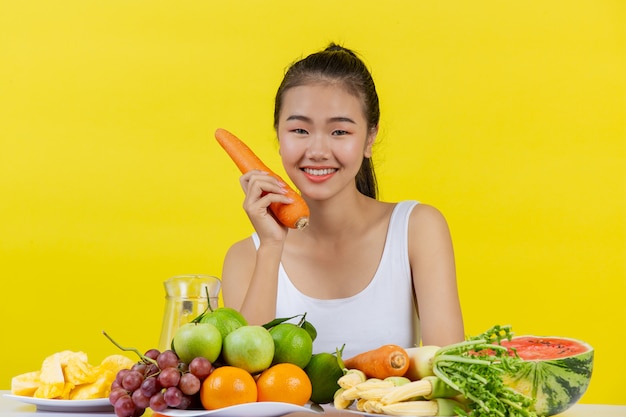 Una mujer asiática vistiendo una camiseta blanca. Sostenga las zanahorias con su mano derecha y en la mesa hay muchas frutas.