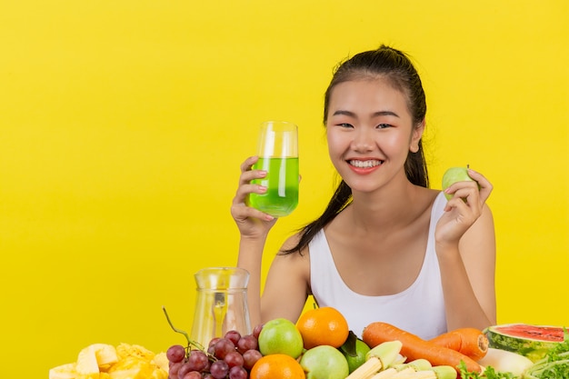 Una mujer asiática vistiendo una camiseta blanca. Sosteniendo un vaso de jugo de manzana con la mano derecha Sosteniendo una manzana con la mano izquierda Y la mesa está llena de varios tipos de frutas.