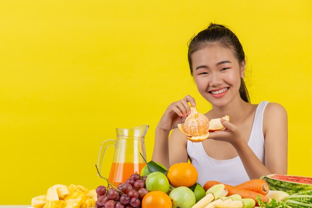 Una mujer asiática vistiendo una camiseta blanca. Soy piel de naranja y la mesa está llena de varios tipos de frutas.