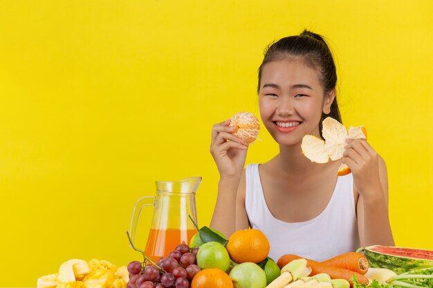 Una mujer asiática vistiendo una camiseta blanca. Soy piel de naranja y la mesa está llena de varios tipos de frutas.