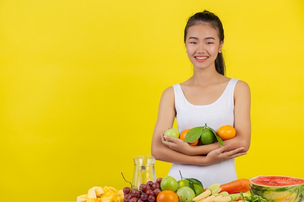 Una mujer asiática vistiendo una camiseta blanca. Usa ambos brazos para sostener varias frutas. Y todavía queda sobre la mesa.