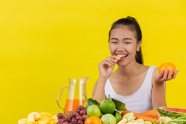 Una mujer asiática vistiendo una camiseta sin mangas blanca comiendo naranja y la mesa está llena de varias frutas.