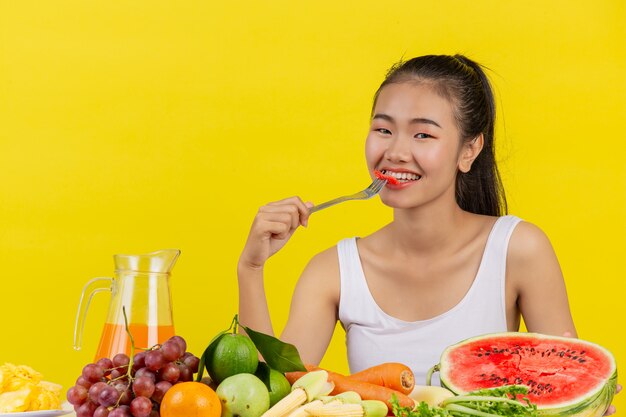 Una mujer asiática vistiendo una camiseta sin mangas blanca comiendo sandía y la mesa está llena de varias frutas.