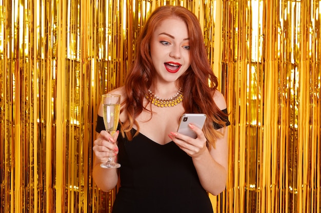 Mujer asombrada con labios rojos y boca ampliamente abierta, mirando con expresión facial de sorpresa en su dispositivo, chica con vestido negro, posando aislada sobre una pared decorada con oropel dorado.