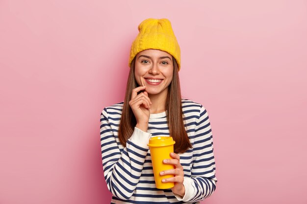 Una mujer de aspecto agradable adora el café, sostiene una taza de comida para llevar, sonríe agradablemente, usa un sombrero amarillo y un suéter a rayas, posa sobre una pared rosa, disfruta del olor